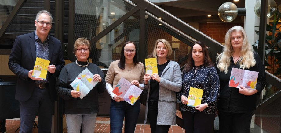 Gruppenbild der Gleichstellungsbeauftragten Inge Keller, Nicole Kruppenbacher, Isabelle Stähle, Sandra Hubert und Marita Rothmann mit Landrat Dietmar Seefeldt vor einem Treppenaufgang im Foyer des Kreishauses der Südlichen Weinstraße in Landau. Alle halten ein Programmhaft der Gleichstellungswochen "Brot und Rosen" 2026 in Händen.