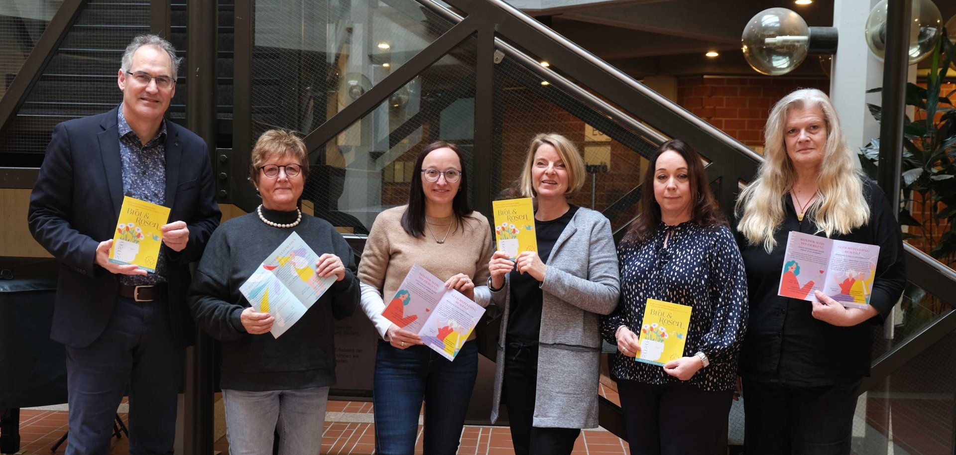 Gruppenbild der Gleichstellungsbeauftragten Inge Keller, Nicole Kruppenbacher, Isabelle Stähle, Sandra Hubert und Marita Rothmann mit Landrat Dietmar Seefeldt vor einem Treppenaufgang im Foyer des Kreishauses der Südlichen Weinstraße in Landau. Alle halten ein Programmhaft der Gleichstellungswochen "Brot und Rosen" 2026 in Händen.