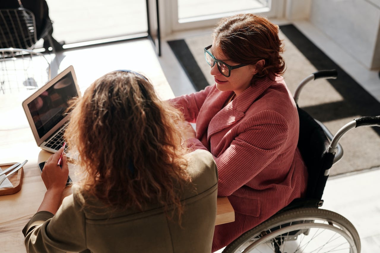 Im Landkreis SÜW kümmert sich ein Beauftragter um die Belange von Menschen mit Behinderungen. Foto: Pexels Zwei Frauen, davon eine im Rollstuhl, sitzen an einem Tisch vor einem Laptop. Foto: Pexels