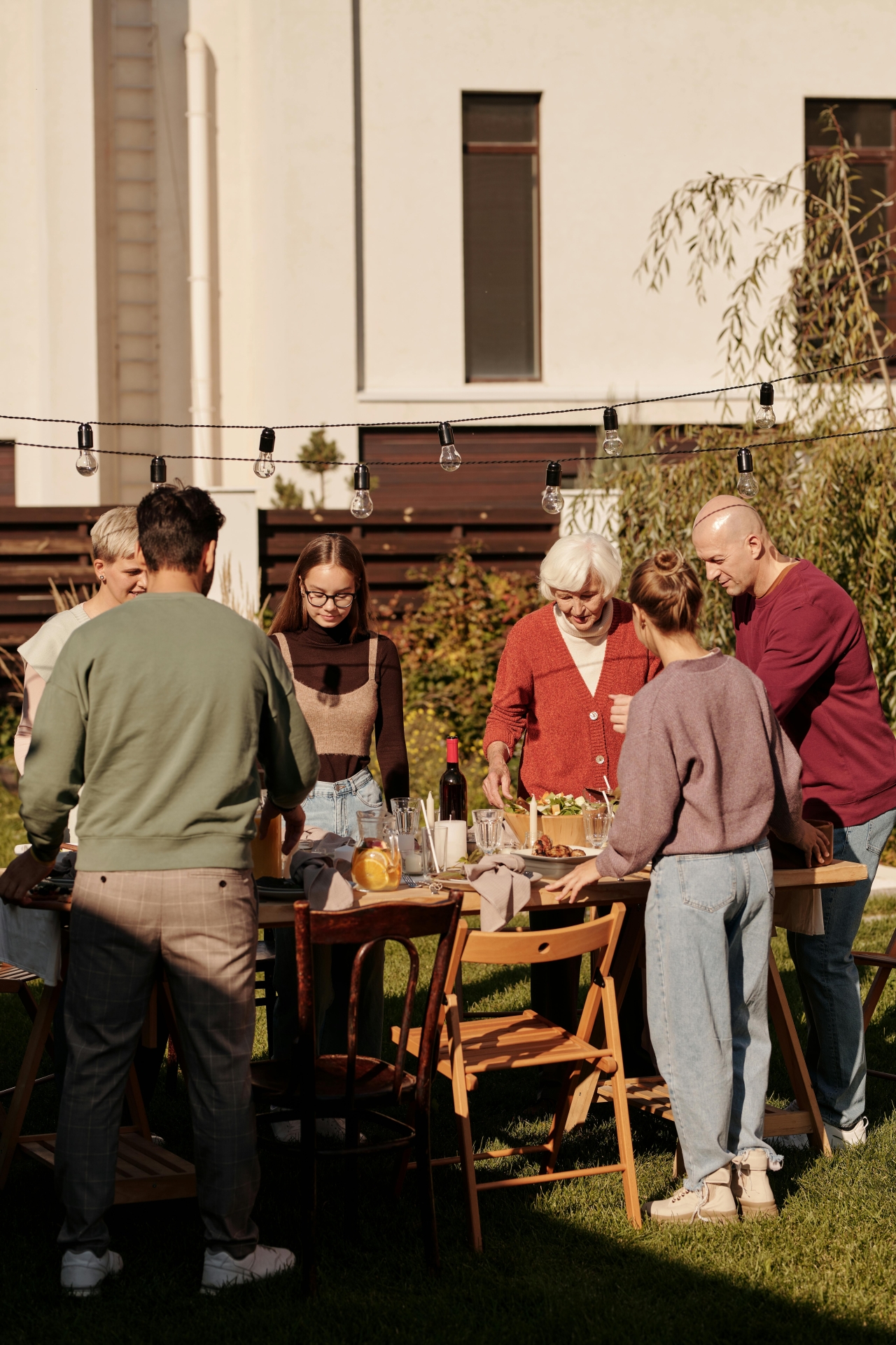 Nachbarschaftstreffen im Garten: Mehrere Menschen verschiedener Generationen stehen gemeinsam um einen gedeckten Tisch im Freien und verbringen Zeit miteinander. Über dem Tisch hängen Lichterketten, die eine gemütliche Atmosphäre schaffen.