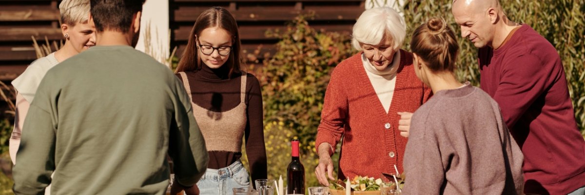Nachbarschaftsglück. Symbolfoto: Pexels Nachbarschaftstreffen im Garten: Mehrere Menschen verschiedener Generationen stehen gemeinsam um einen gedeckten Tisch im Freien und verbringen Zeit miteinander. Über dem Tisch hängen Lichterketten, die eine gemütliche Atmosphäre schaffen.
