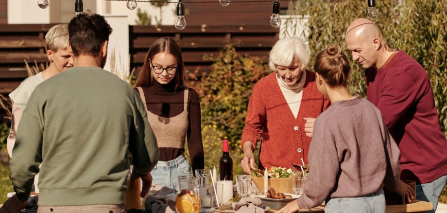 Nachbarschaftsglück. Symbolfoto: Pexels Nachbarschaftstreffen im Garten: Mehrere Menschen verschiedener Generationen stehen gemeinsam um einen gedeckten Tisch im Freien und verbringen Zeit miteinander. Über dem Tisch hängen Lichterketten, die eine gemütliche Atmosphäre schaffen.