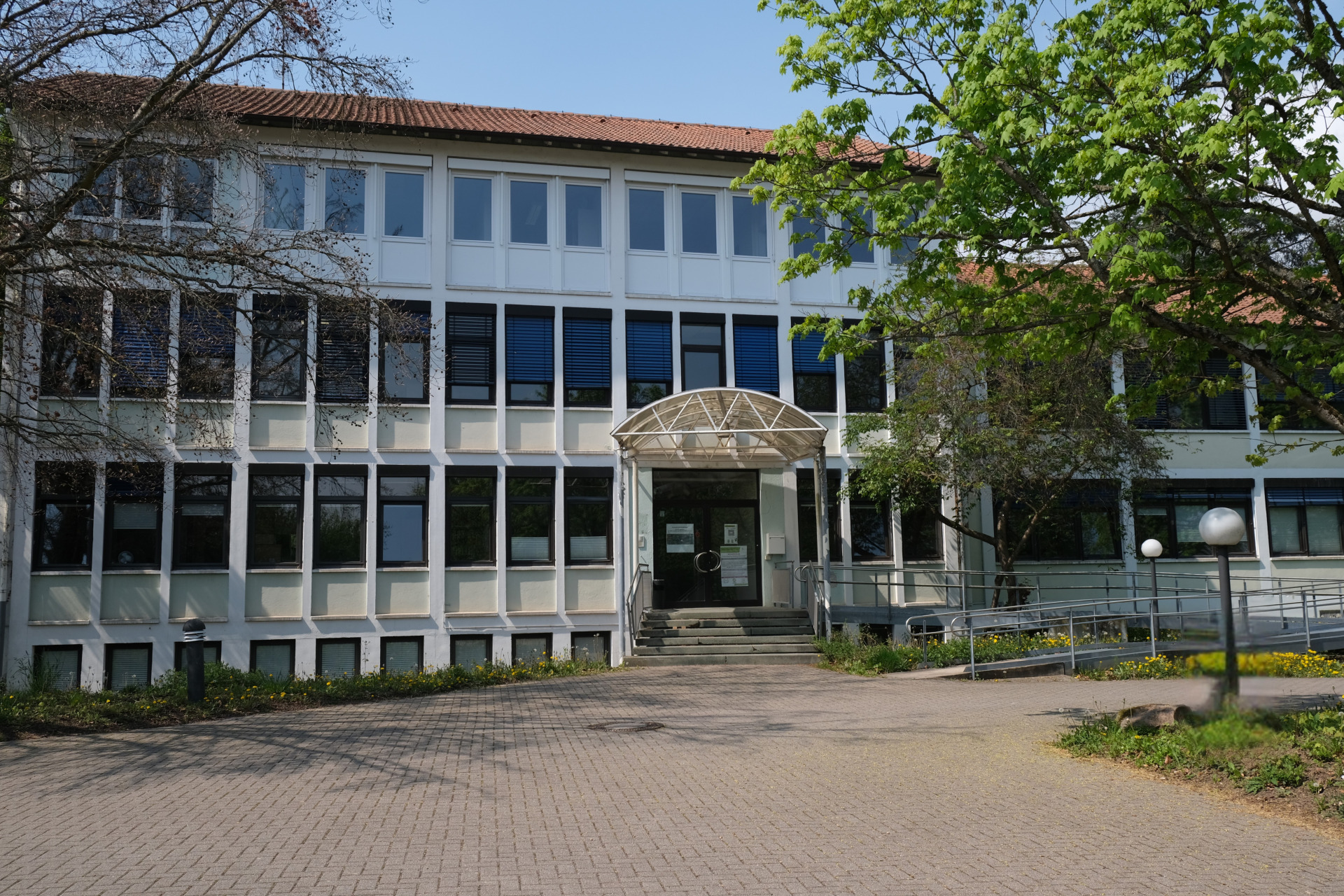 Frontalansicht eines mehrgeschossigen Verwaltungsgebäudes mit heller Fassade und rotem Ziegeldach. In der Mitte führt eine Treppe zu einem überdachten Haupteingang mit Glasvordach.