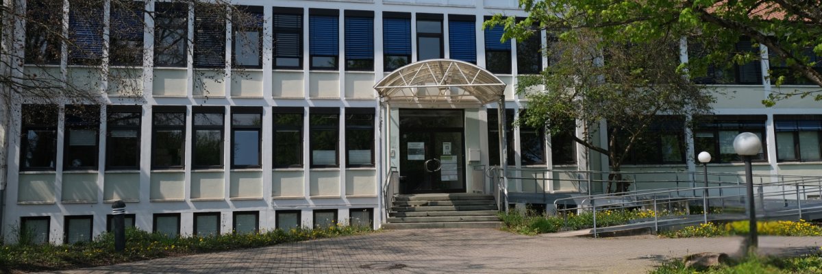 Frontalansicht eines mehrgeschossigen Verwaltungsgebäudes mit heller Fassade und rotem Ziegeldach. In der Mitte führt eine Treppe zu einem überdachten Haupteingang mit Glasvordach.