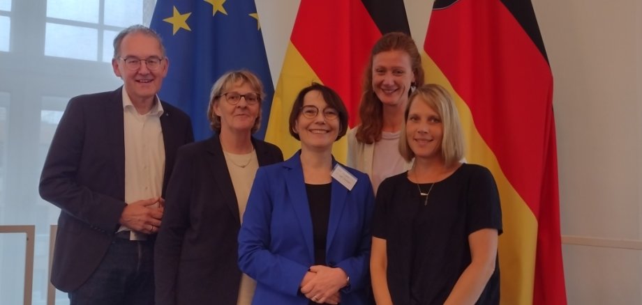 Festakt in der Staatskanzlei Rheinland-Pfalz: Seit zehn Jahren sind die Fachkräfte Gemeindeschwester plus aktiv – der Landkreis SÜW war von Beginn an dabei. Foto: Fünf Personen stehen gemeinsam vor den Flaggen der Europäischen Union, Deutschlands und von Rheinland-Pfalz. Die Gruppe posiert lächelnd für ein Foto bei einer offiziellen Veranstaltung.